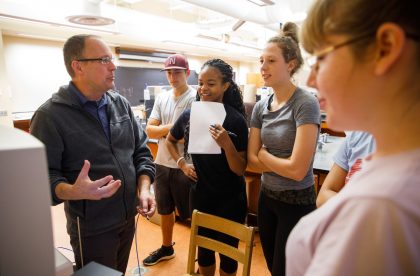 Faculty member conversing with a group of students in a classroom setting.