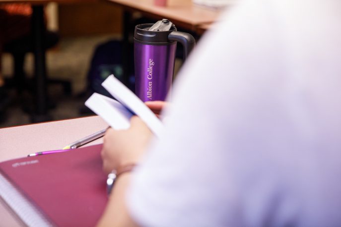 Student holding a book at a desk in a classroom setting.