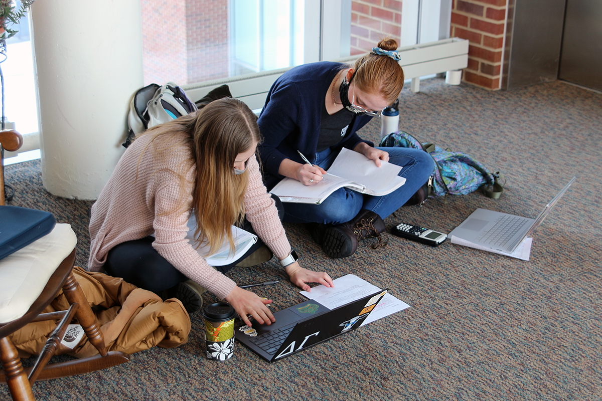 Two students wearing masks working on the floor together working on laptop computers