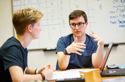 Two students conversing in front of a whiteboard.