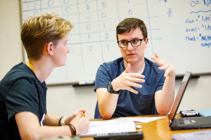Two students conversing in front of a whiteboard.