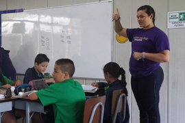 Three children sit at a table with books in front of them. A female teacher stands behind them, gesturing with her hands. A whiteboard is behind them on the wall.