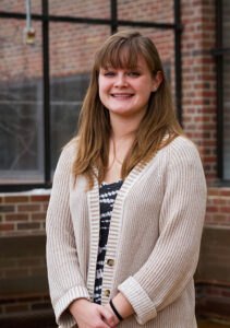 A female student wearing a cream sweater standing in front of a window, smiling.