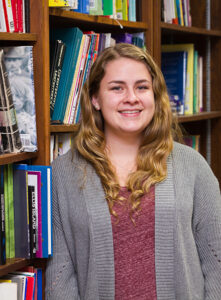 A female student wearing a burgundy shirt and gray cardigan standing in front of a bookcase.