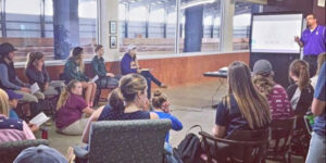 Students sitting in chairs and on the floor facing the front of a room, where a teacher is giving a PowerPoint presentation.