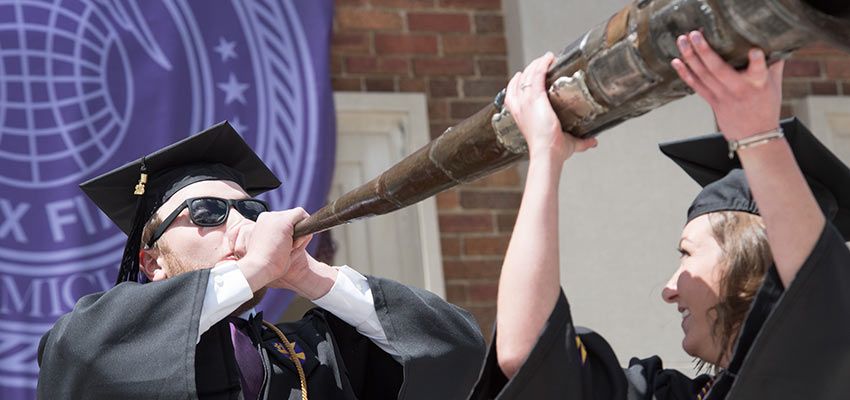 A student blowing into a large instrument while another student holds it up.