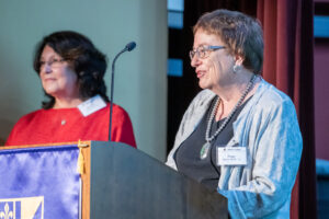A photograph of Peggy Sindt standing behind a podium.