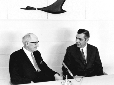 E.T.S. Walton wears a suit and glasses. He is talking to another man in a suit and tie as they sit at a table with a microphone on it.