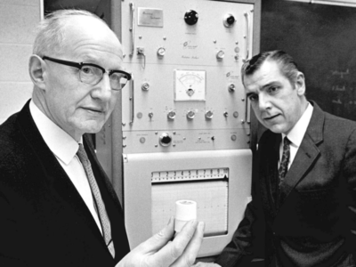 E.T.S. Walton is holding a small white cylinder. A man in the background is looking at the cylinder/ A scientific looking machine is behind them both.