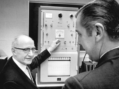 E.T.S. Walton is turning the knob of a scientific looking machine while he talks to a man in the foreground.