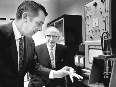 E.T.S. Walton smiles as the man in the foreground examines a small white cylinder.