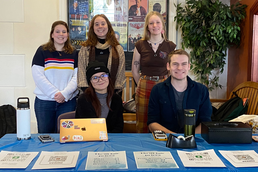 Two students sit at table with a laptop and calculators. Three students stand behind them. 