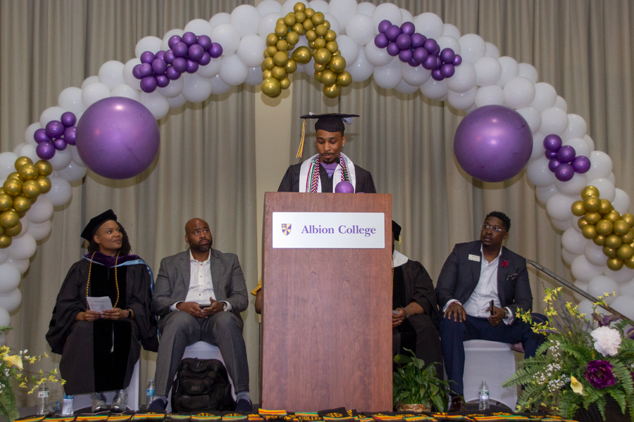 A student wearing academic regalia addresses an audience of graduates from a podium.