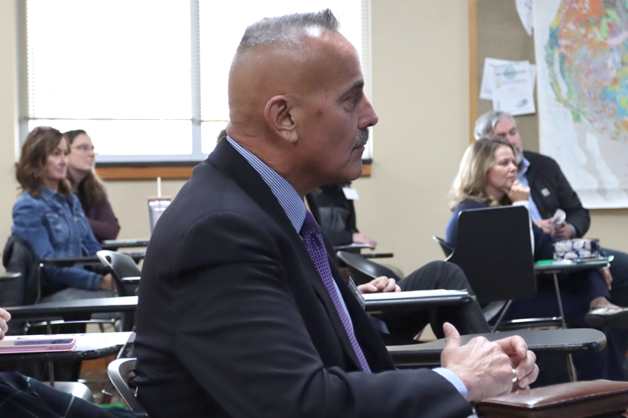 Bill is sitting at a desk, intently watching the presentation.