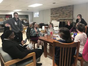 People laughing and standing around a wooden table with some snacks and papers